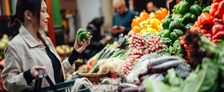 An adult carrying a shopping basket picks up and inspects a green pepper from a selection of other vegetables and fruit.