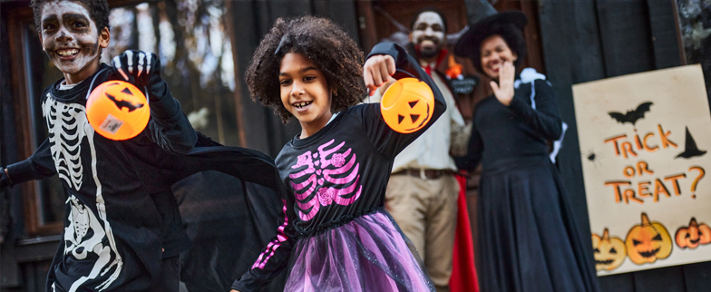 Two kids trick or treating in Halloween costumes holding up their pumpkin candy bags with their parents behind them