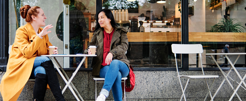 Two friends sitting outside at a coffee shop talking while drinking a cup of coffee. 